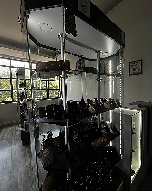 Vitrina de cristal iluminada con joyería de piercing en soportes negros; al fondo, el ventanal con árboles y los estantes de supply